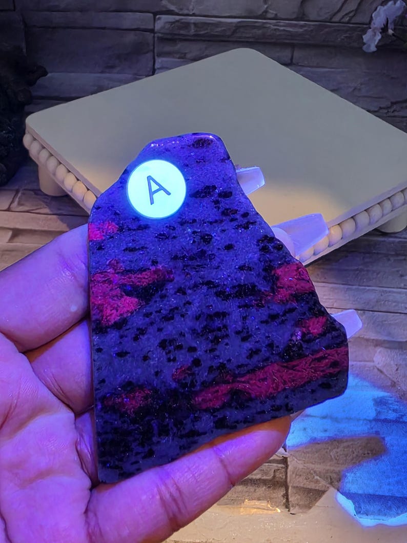 Op de afbeelding: Een platte, onregelmatig gevormde plaat van mineraalsteen met een donkerpaarse en zwarte basis met levendige, gloeiende magentakleurige vlekken. Een ronde witte sticker met de letter A is bevestigd op het bovenste deel van het stenen oppervlak.