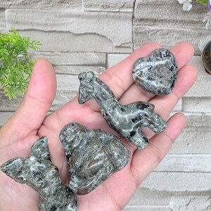 May include: A collection of carved stone figurines in various shapes, including a heart, a giraffe, a seated figure, and a butterfly. The figurines are primarily gray with black and brown accents, and are held in a hand.