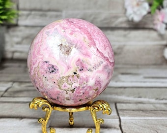 May include: A pink sphere with white and gold veining sits on a gold-colored decorative stand. The sphere has a polished surface and a marbled appearance. The stand has a detailed, ornate design with three legs.