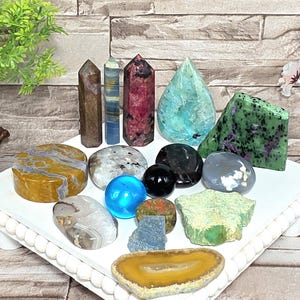 May include: A collection of various polished and raw mineral specimens including three vertical crystal points, a teardrop-shaped blue stone, a triangular green and black stone, a bright blue sphere, a black sphere, and several flat, rounded stones.