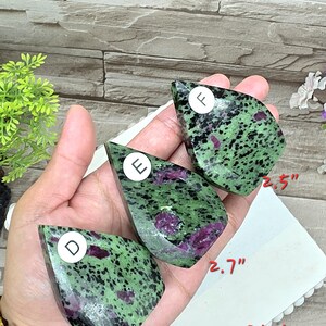 May include: Three teardrop-shaped polished stones featuring a green and black speckled pattern with embedded magenta-colored inclusions. Each stone is labeled with a white circular sticker marked D, E, or F, with visible measurements of 2.5 inches and 2.7 inches.