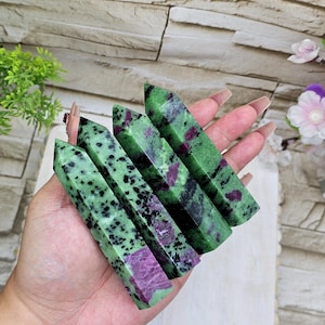 May include: Four green and red zoisite crystal wands are held in a hand. The crystals have a polished, rectangular shape with a mix of green and black speckles and patches of deep red. The background is a light-colored brick wall.