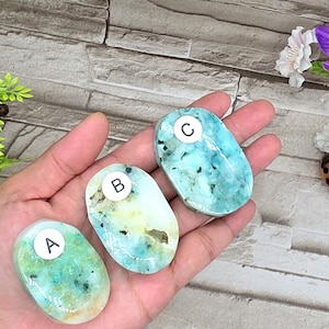 May include: Three polished, oval-shaped gemstones in shades of turquoise, green, and white are displayed. Each stone has a unique pattern and is labeled with a letter. The gemstones are held in a hand, showcasing their natural beauty and variations.