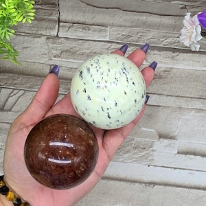 May include: Two spherical gemstones. One is a light green color with black speckles, and the other is a reddish-brown color. The gemstones are smooth and polished, and appear to be held in a hand. The background is a light gray brick wall.