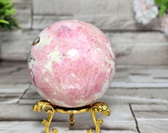 May include: A pink and white sphere, possibly a gemstone, sits on a decorative gold-colored stand. The sphere has a marbled appearance with varying shades of pink and white. The stand has an ornate, scrolled design.