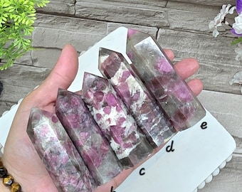 May include: Five polished crystal points arranged in a row, each featuring a mix of translucent smoky quartz and vibrant magenta-pink inclusions. The crystals are labeled with the letters a, b, c, d, and e written on the white surface below them.