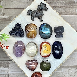 May include: A collection of polished gemstone carvings including a horse, a Buddha, a cat, a heart, an apple, and a skull, alongside various oval and rounded stones in shades of purple, brown, blue, clear, red, and green on a white tray.