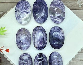 May include: Nine oval-shaped cabochons made of purple and white charoite stone. Each piece features unique swirling patterns of deep violet, lavender, and white mineral inclusions with a polished, glossy surface finish arranged in a grid.
