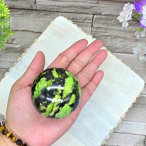 May include: A polished sphere with a vibrant green and black pattern. The gemstone is held in an open hand, showcasing its smooth, rounded shape. The sphere's colors are reminiscent of a watermelon. The background is a light-colored wooden surface.