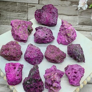 May include: A collection of vibrant, rough-textured, magenta-colored mineral specimens. The stones vary in size and shape, with a crystalline appearance. They are arranged on a white surface with a beaded edge, set against a neutral background.