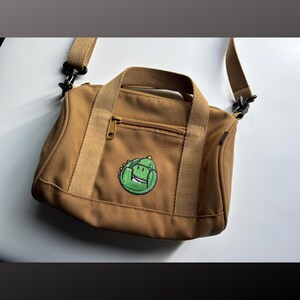 May include: A tan canvas crossbody bag featuring a small, circular embroidered patch of a green dinosaur on the front pocket. The bag includes a zippered front compartment, two top handles, and an adjustable shoulder strap with black hardware.