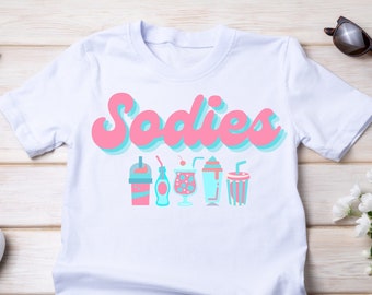 Peut inclure: T-shirt blanc avec le mot "Sodies" en rose et bleu, et une rangée d'illustrations de boissons stylisées en rose et bleu. Des lunettes de soleil et des chaussures blanches sont visibles.
