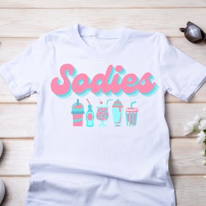 Può includere: Maglietta bianca con la scritta "Sodies" in rosa e blu, e una fila di illustrazioni stilizzate di bevande in rosa e blu. Occhiali da sole e scarpe bianche sono visibili.