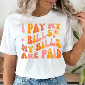Może przedstawiać: Biała koszulka z napisem "I PAY MY BILLS, MY BILLS ARE PAID" w pomarańczowych, różowych i żółtych literach bąbelkowych. Koszulka ma retro design z kwiatowymi akcentami.