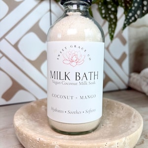 May include: A clear glass bottle filled with off-white powder, topped with a black cap. The white label reads Sweet Grace Co, Milk Bath, Vegan Coconut Milk Soak, Coconut + Mango, Hydrates, Soothes, Softens, with a lotus flower graphic.