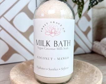 May include: A clear glass bottle filled with off-white powder, topped with a black cap. The white label reads Sweet Grace Co, Milk Bath, Vegan Coconut Milk Soak, Coconut + Mango, Hydrates, Soothes, Softens, with a lotus flower graphic.