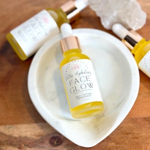 May include: A glass bottle of yellow face glow serum with a white label that reads "Sweet Grace Ultra Hydrating Face Glow Organic | Cold Pressed Hydrating Facial Oil 1 fl oz | 45 ml"