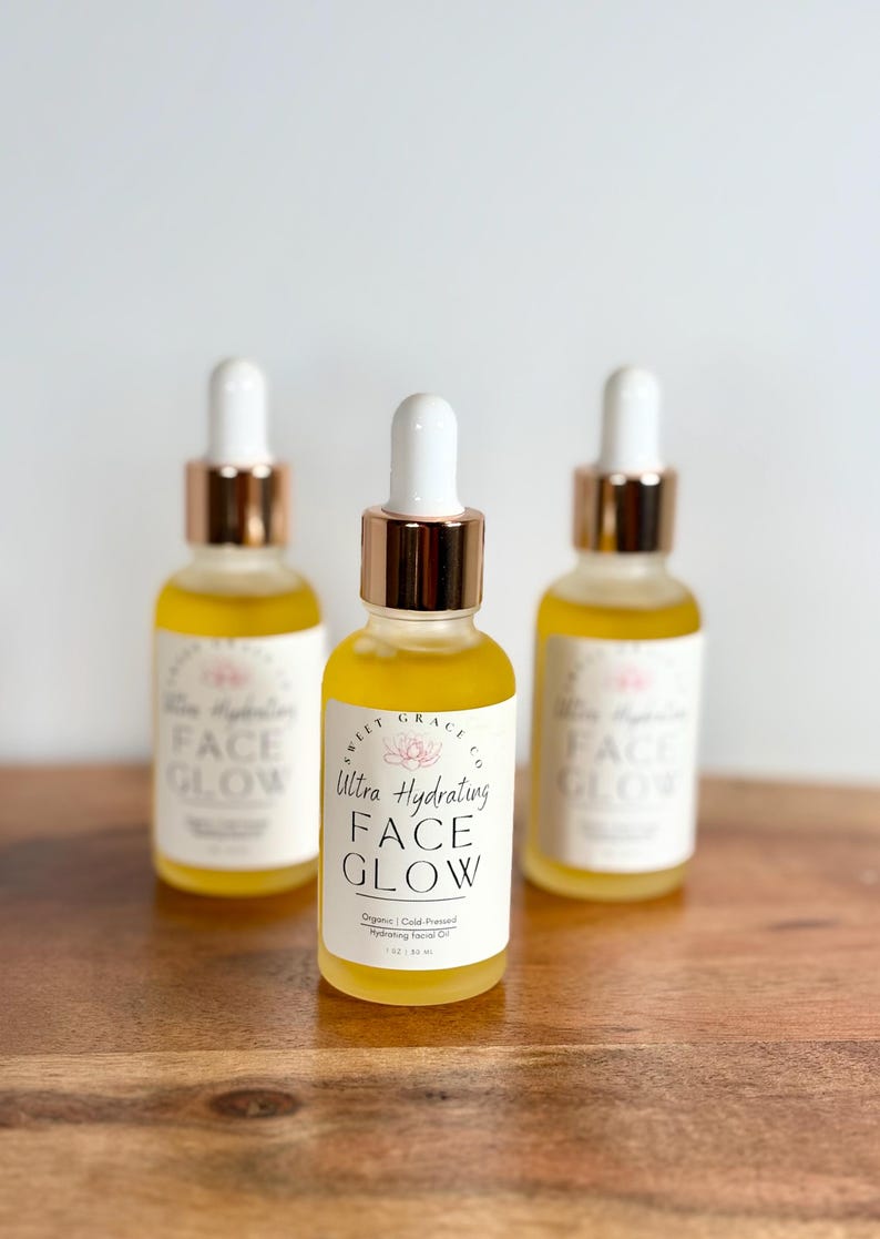 May include: Three amber glass bottles with gold caps and droppers filled with a yellow liquid. The bottles are labelled "Sweet Grace Co. Ultra Hydrating Face Glow. Organic | Cold Pressed Hydrating Facial Oil 1 fl oz / 30 ml."