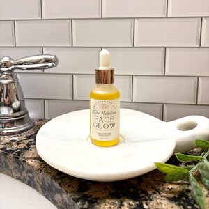 May include: A small, clear glass bottle with a gold cap and a dropper. The bottle is filled with a yellow liquid and has a label that reads "Ultra Hydrating Face Glow". The bottle is sitting on a white marble dish.