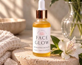 May include: A clear glass dropper bottle containing golden yellow facial oil with a white label reading FACE GLOW, HYDRATING FACIAL OIL, Organic, Vegan, Crystal-Infused, and a logo with a lotus flower, topped with a white and gold dropper cap.