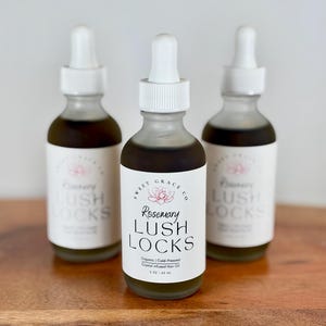 May include: Three glass bottles with white dropper tops and labels. The center bottle is labeled "Rosemary Lush Locks Organic Cold-Pressed Crystal Infused Hair Oil 2 oz / 60 ml". The other two bottles are labeled "Lush Locks".
