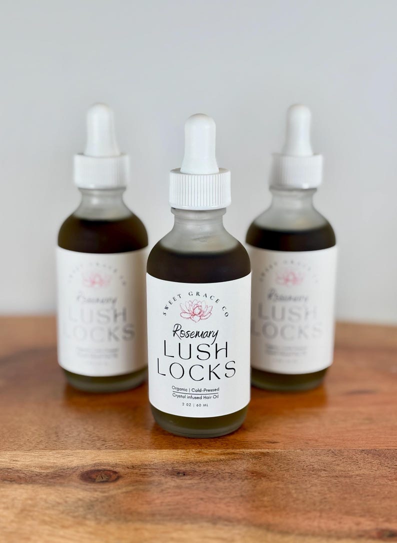 May include: Three glass bottles with white dropper tops and labels. The center bottle is labeled "Rosemary Lush Locks Organic Cold-Pressed Crystal Infused Hair Oil 2 oz / 60 ml". The other two bottles are labeled "Lush Locks".