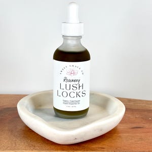 May include: A clear glass bottle with a white dropper top filled with a dark green liquid. The bottle is labeled "Rosemary Lush Locks Organic Cold Pressed Crystal Infused Hair Oil 2 oz / 60 ml". The bottle is sitting on a white heart-shaped dish.