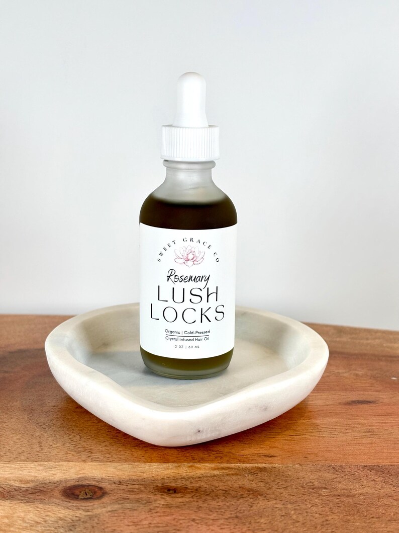 May include: A clear glass bottle with a white dropper top filled with a dark green liquid. The bottle is labeled "Rosemary Lush Locks Organic Cold Pressed Crystal Infused Hair Oil 2 oz / 60 ml". The bottle is sitting on a white heart-shaped dish.
