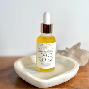 May include: A clear glass bottle with a gold cap and a dropper top. The bottle contains a yellow liquid and has a white label with the text "Sweet Grace Co. Ultra Hydrating Face Glow Organic | Cold-Pressed Hydrating Facial Oil 1 fl oz | 30 ml".