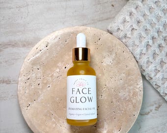 May include: A clear glass dropper bottle containing yellow facial oil with a white label that reads Sweet Grace Co Face Glow Hydrating Facial Oil Organic Vegan Crystal-Infused. The bottle has a gold collar and a white dropper top.