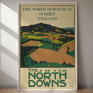 May include: A framed vintage poster advertising The North Downs Way in Surrey, England. The artwork depicts a landscape of rolling hills and fields in shades of green, brown, and yellow. The poster's text reads "The North Downs Way Surrey England" in a vintage font.