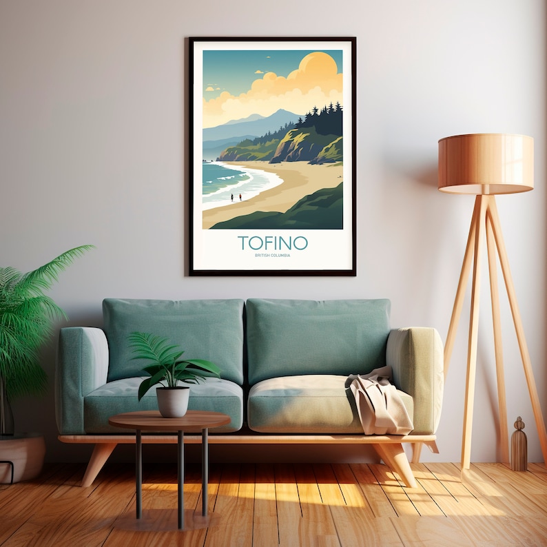 Op de afbeelding: Een ingelijste kunstprint met een kustlandschap met de tekst "TOFINO BRITISH COLUMBIA". Het kunstwerk toont een strand, oceaan, bergen en een ondergaande zon in tinten blauw, groen en geel. Twee figuren lopen op het strand.