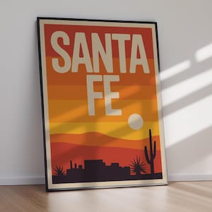 May include: Framed poster with the words "SANTA FE" in large, off-white letters. The background features a sunset gradient of orange and yellow hues, with a silhouette of a desert landscape, including cacti and buildings.