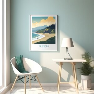 Op de afbeelding: Een ingelijste kunstafdruk met een kustlandschap met een strand, zee en bergen onder een bewolkte hemel. De afdruk heeft het woord "TOFINO" en "British Columbia" in een witte lijst. De afdruk hangt aan een lichtblauwe muur.
