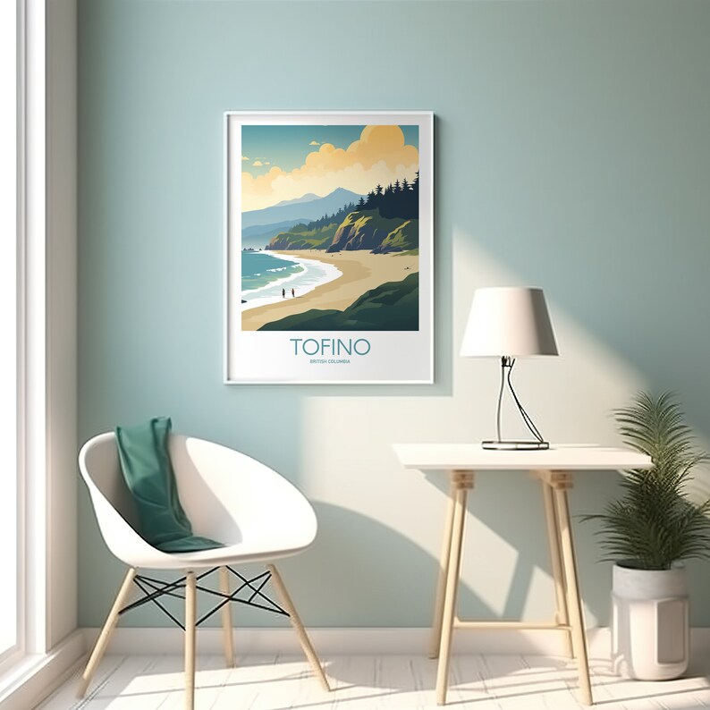 Op de afbeelding: Een ingelijste kunstafdruk met een kustlandschap met een strand, zee en bergen onder een bewolkte hemel. De afdruk heeft het woord "TOFINO" en "British Columbia" in een witte lijst. De afdruk hangt aan een lichtblauwe muur.