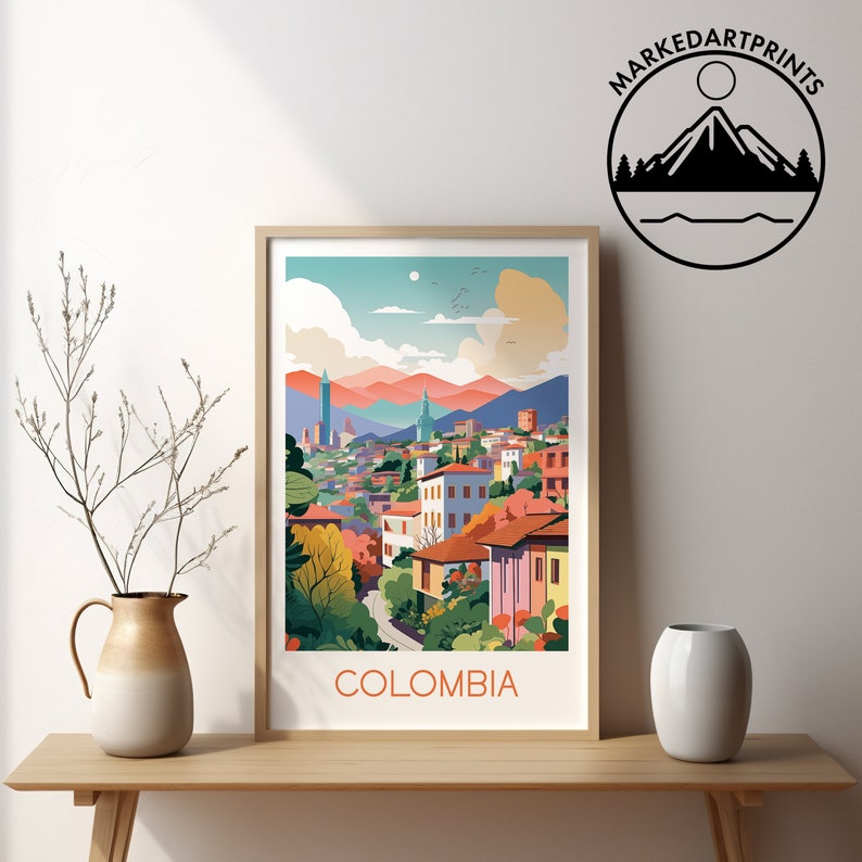 May include: A framed art print featuring a colorful illustration of a cityscape with buildings, mountains, and a blue sky. The word "COLOMBIA" is printed at the bottom. The print is displayed on a wooden surface with a vase and decorative branches.