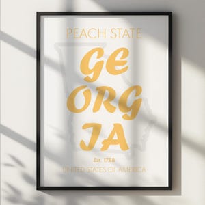 May include: Framed art print featuring the state of Georgia. The words "GEORGIA" are in a bold, yellow font, with the outline of the state in the background. The text "PEACH STATE", "Est. 1788", and "UNITED STATES OF AMERICA" are also included.