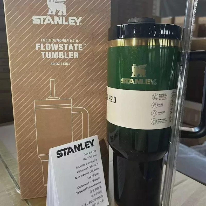 May include: A green and black Stanley Flowstate tumbler with a handle and straw. The tumbler has gold accents and the Stanley logo. The box reads "The Quencher H2.0 Flowstate Tumbler 40 oz / 1.18L". A care and use booklet is also visible.