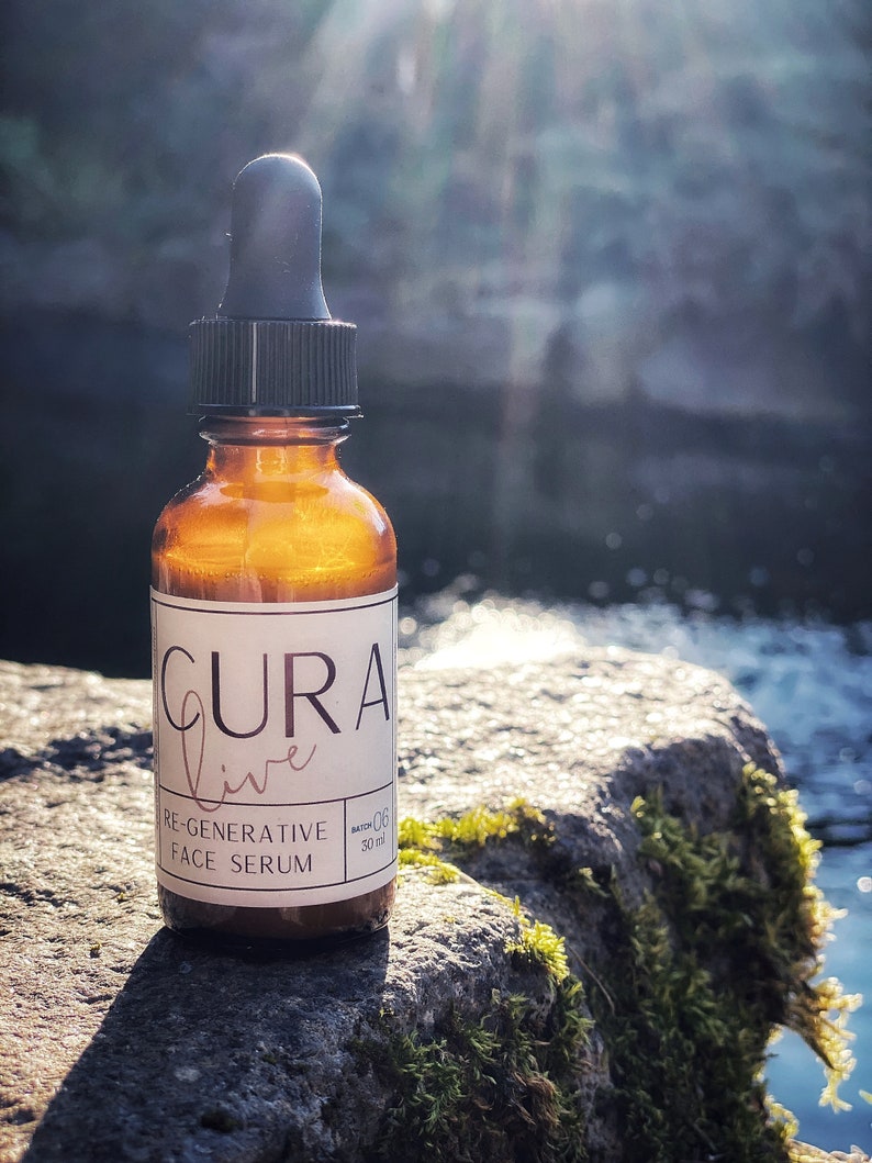 May include: A brown glass bottle with a dropper top and a white label that reads "CURA live RE-GENERATIVE FACE SERUM" and "BATCH 06 30 ml". The bottle is sitting on a mossy rock.