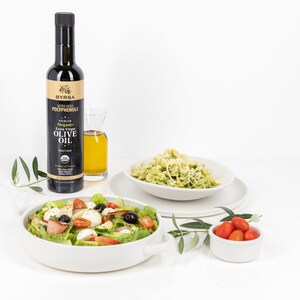 May include: A tall dark bottle of Byrsa organic extra virgin olive oil with a gold label, next to a small glass carafe of oil, a white bowl of salad with tomatoes and mozzarella, a white bowl of pasta, and a small bowl of cherry tomatoes.