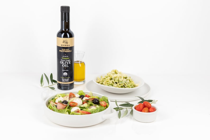 May include: A tall dark bottle of Byrsa organic extra virgin olive oil with a gold label, next to a small glass carafe of oil, a white bowl of salad with tomatoes and mozzarella, a white bowl of pasta, and a small bowl of cherry tomatoes.