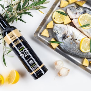 May include: A black bottle of Byrsa organic extra virgin olive oil with a gold label, positioned next to a metal tray containing two whole raw fish, lemon slices, potato chunks, and peppercorns, accompanied by fresh olive branches.