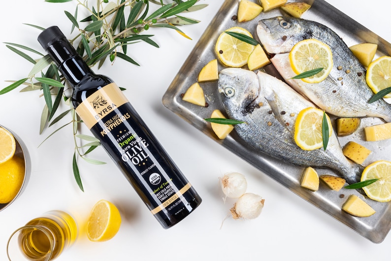 May include: A black bottle of Byrsa organic extra virgin olive oil with a gold label, positioned next to a metal tray containing two whole raw fish, lemon slices, potato chunks, and peppercorns, accompanied by fresh olive branches.