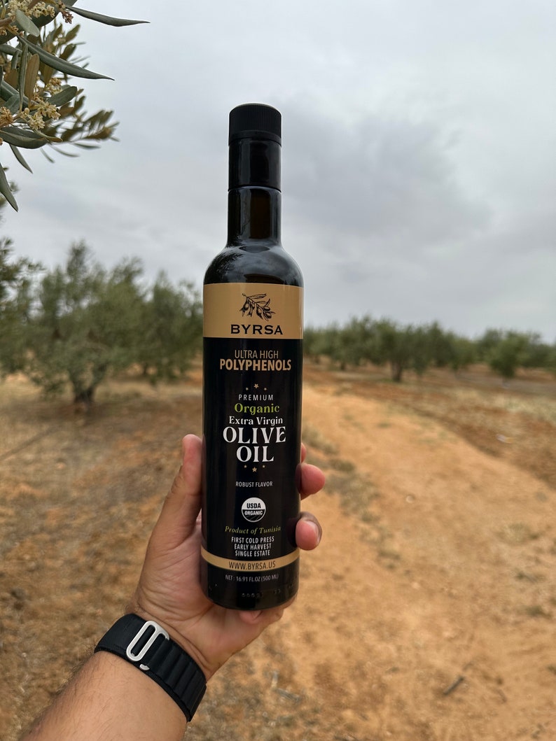 May include: A tall, dark glass bottle of Byrsa organic extra virgin olive oil with a black cap. The label features the text Ultra High Polyphenols, Premium Organic Extra Virgin Olive Oil, Robust Flavor, USDA Organic, Product of Tunisia, and 500 ml.