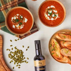 May include: Two bowls of red tomato soup topped with cream and herbs, a plate of toasted bread slices, a bottle of Byrsa organic extra virgin olive oil, a scattering of green pumpkin seeds, and two wooden spoons on a textured white surface.