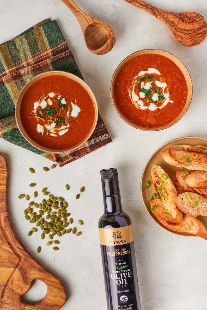 May include: Two bowls of red tomato soup topped with cream and herbs, a plate of toasted bread slices, a bottle of Byrsa organic extra virgin olive oil, a scattering of green pumpkin seeds, and two wooden spoons on a textured white surface.