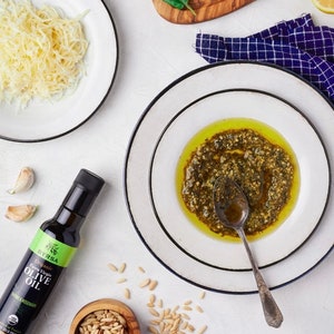 May include: A bottle of Byrsa Organic Extra Virgin Olive Oil with thyme and rosemary, a bowl of pine nuts, a plate of shredded cheese, and a plate containing green pesto sauce with a metal spoon, arranged with fresh basil and lemon slices.