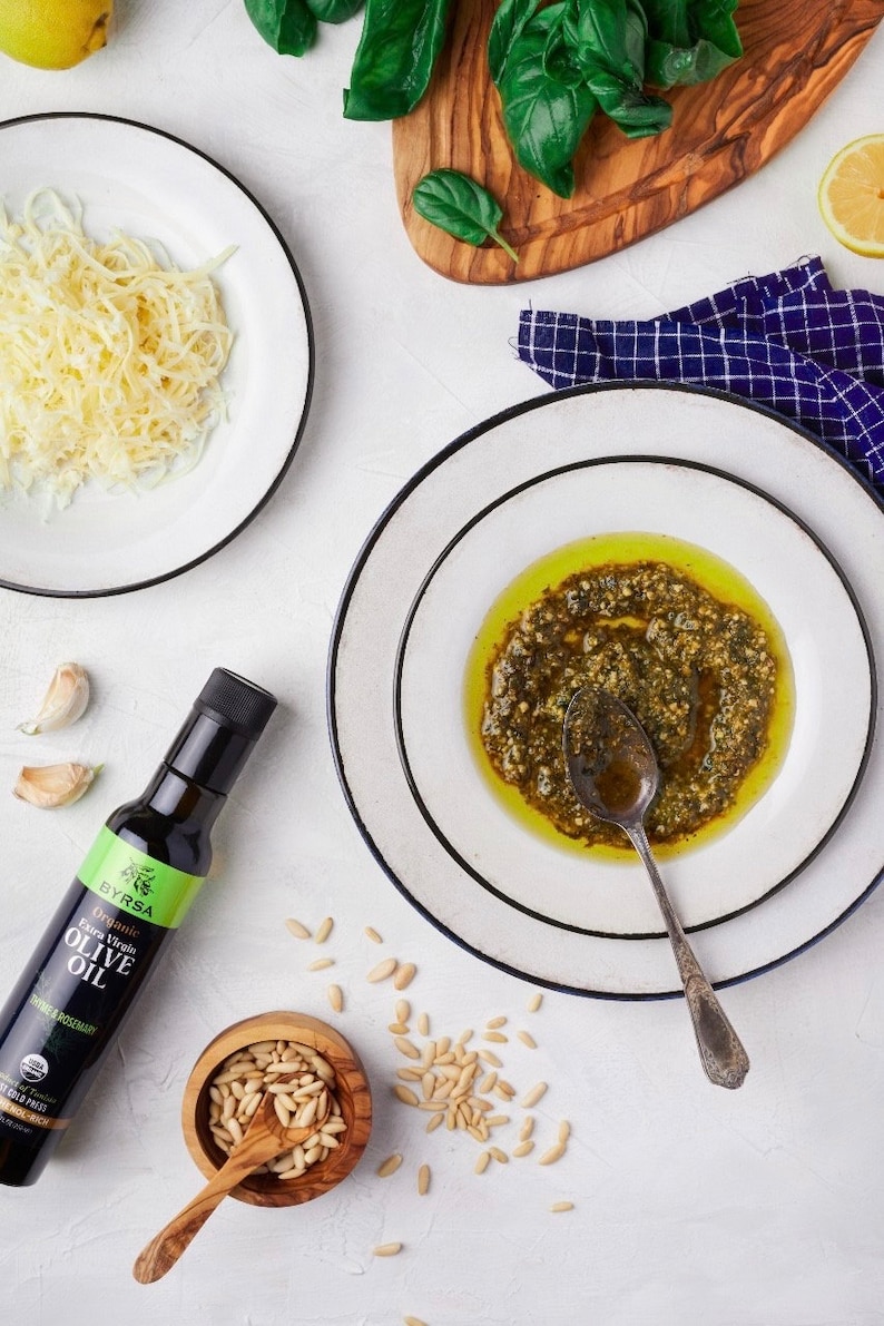 May include: A bottle of Byrsa Organic Extra Virgin Olive Oil with thyme and rosemary, a bowl of pine nuts, a plate of shredded cheese, and a plate containing green pesto sauce with a metal spoon, arranged with fresh basil and lemon slices.