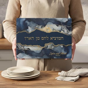May include: A navy blue and gold decorative canvas with Hebrew text and the names "Sylvia and Micah" with the date "10.18.25". The canvas has a watercolor design with gold accents.