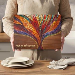 May include: A rectangular wooden cutting board with a colorful mosaic tree design. The tree features red, orange, yellow, and blue hues. The bottom portion of the board has a wood grain pattern with gold text.